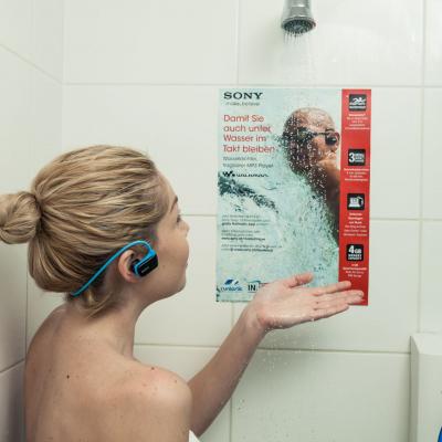 Frau steht im Fitness Studio unter Dusche mit Walkman, an den Kacheln Werbung für wasserdichten Sony Walkman