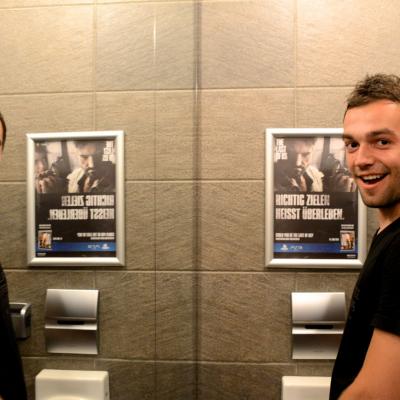 2 Junge Männer in gepflegtem Pissoir im Portrait, auf der Rückwand Werbeposter